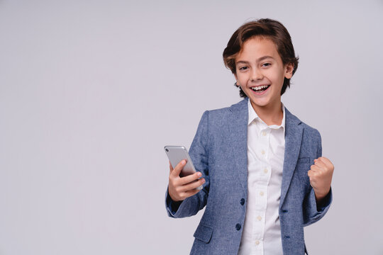 Small cute boy celebrating like a winner while holding mobile phone isolated on white background, childhood, technology, success, cheerful emotion and lifestyle concept