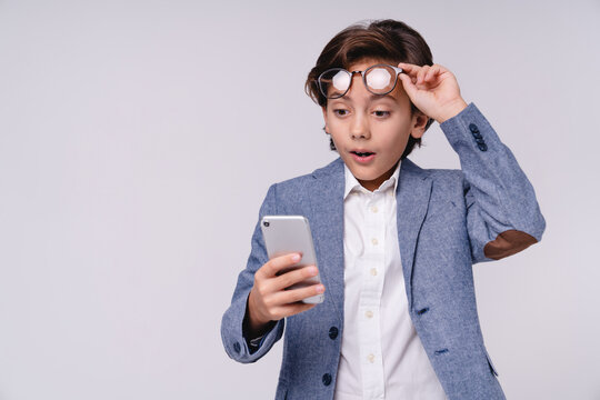 Surprised little boy in formal outfit looking at smartphone with shocked expression isolated on grey background, childhood, technology, communication, emotion and future businessman concept
