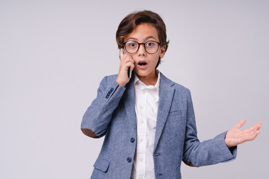 Shocked young caucasian boy in formal clothes talking on phone isolated on grey background, childhood, communication, technology, emotion and future businessman concept