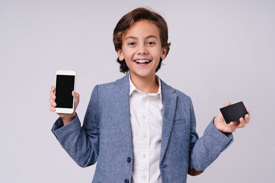 Smiling young boy holding credit card and mobile phone isolated on grey background, childhood, technology, shopping, payment, finance, online purchase and lifestyle concept