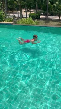 Woman swims in clear pool, making waves as they move. The sun shines brightly on water, reflecting light and creating a playful look. It is a warm day.