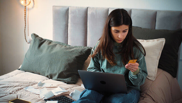 Young Latina woman shopping online from her bed despite having significant debts, highlighting the contrast between financial pressure and consumer spending.