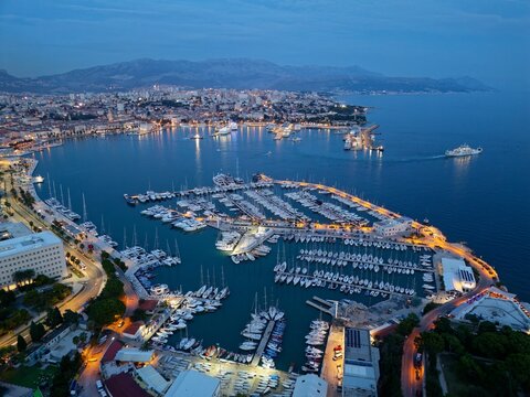 Majestic twilight aerial view of Split, Croatia, where illuminated harbors, glowing marinas, and red-roofed coastal stone buildings meet the calm blue Adriatic Sea beneath distant alpine ridges.