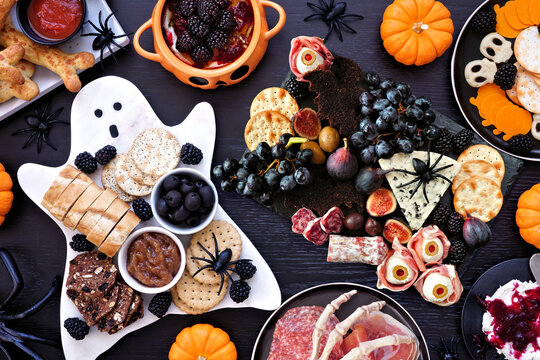 Halloween theme grazing table scene against a black wood background. Spooky charcuterie boards of meat and cheese appetizers. Top view.