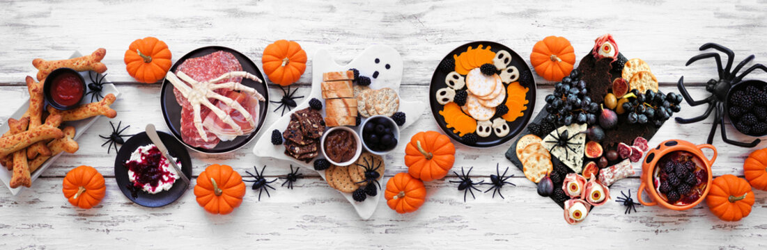 Halloween theme grazing table scene against a white wood banner background. Spooky charcuterie boards of meat and cheese appetizers. Top down view.