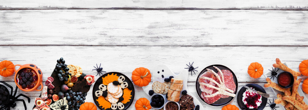Halloween theme grazing bottom border against a white wood banner background. Spooky charcuterie boards of meat and cheese appetizers. Top view with copy space.