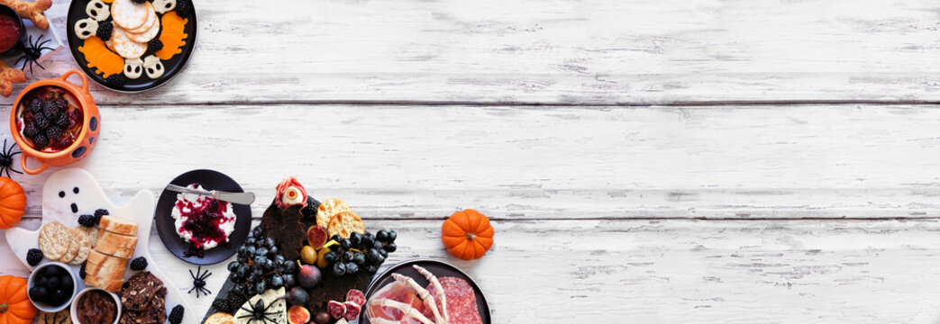 Halloween theme grazing corner border against a white wood banner background. Spooky charcuterie boards of meat and cheese appetizers. Overhead view with copy space.