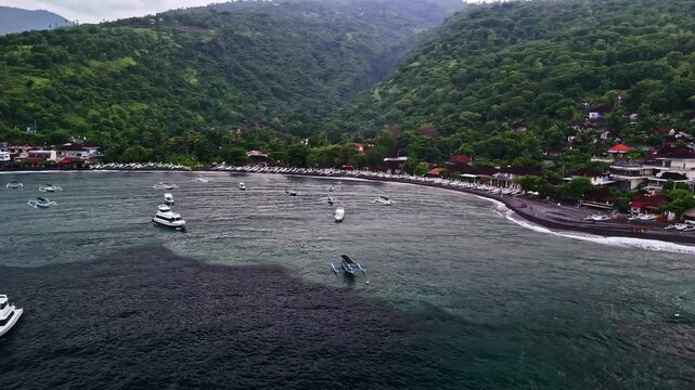 Aerial view of Amed Beach featuring traditional boats on the water, dark volcanic sand coastline, and lush green hills in Bali, Indonesia.