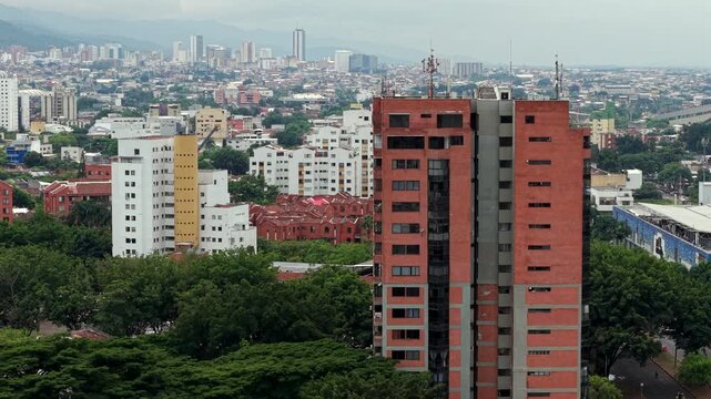 Sweeping aerial footage of Cali&rsquo;s Calle Novena featuring modern and historic buildings, tall red-brick towers, and abundant city greenery under soft daylight. Ideal for urban landscape themes.