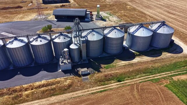Closer panoramic drone fly of metal grain silos stand in a row beside harvested fields showing rural agricultural storage infrastructure.