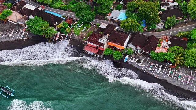 Aerial view of Amed Beach featuring traditional outrigger boats on the black sand shore with turquoise waves and coastal buildings in Bali, Indonesia.