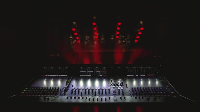 Dark stage setting with professional audio mixing console in foreground, illuminated faders buttons, red stage lights in background creating dramatic atmosphere, shallow depth of field, ideal for..
