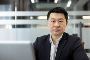Fototapeta premium Asian businessman confidently looking at camera while sitting at his desk with a laptop, reflecting professionalism, corporate success, and focused determination in a contemporary office environment