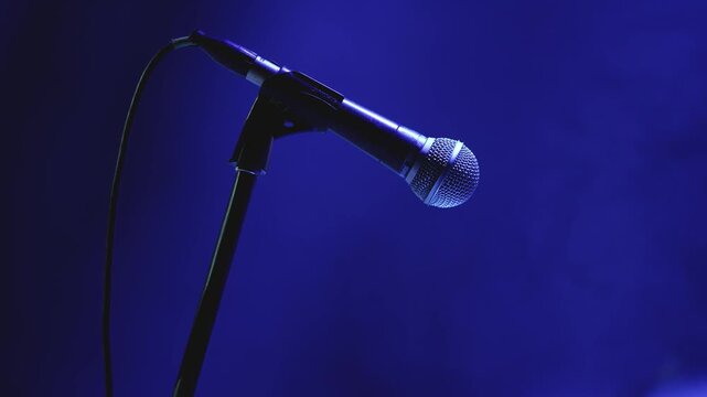 Close up of microphone on stand bathed in deep blue stage light. Soft bokeh background suggests live performance setting. Shallow depth of field highlights mic grille texture