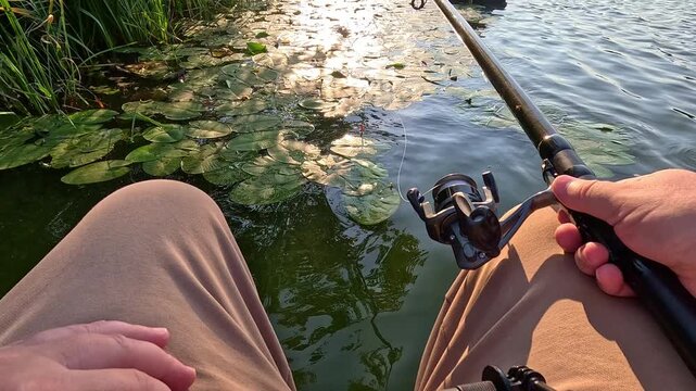 First person view of a hand holding a fishing rod over the calm water of a lake covered with water lilies, sunlight reflecting on the surface near a wooden dock in daylight.
