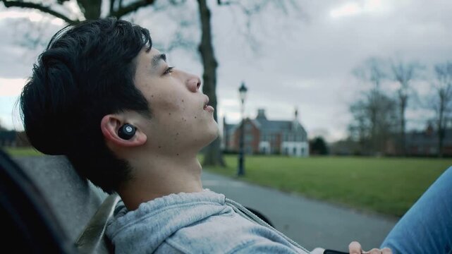 Leaning back, Asian male wearing hoodie reclining on campus bench, gazing up with phone and earbud
