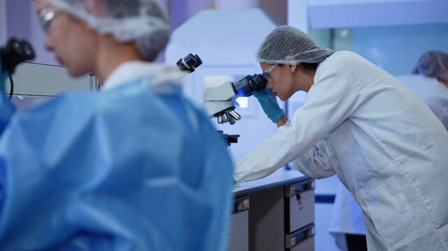 Multiple researcher lean over microscope in a busy laboratory, wearing hairnet and safety glasses while conducting scientific analysis and sample examination together