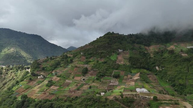 Drone flies forward toward cultivated hillside farms surrounded by lush mountains and low clouds in Zunil, Quetzaltenango, Guatemala.