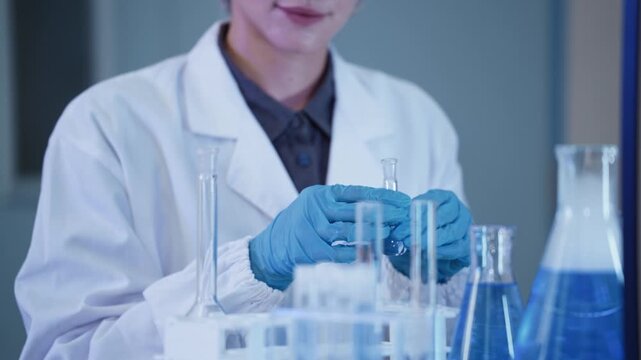 Close up of a gloved hand pouring liquid into a graduated cylinder beside flask filled with blue liquid, representing chemistry research and laboratory experiment concept