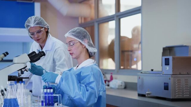 A researcher holds up a test tube with blue liquid while a colleague seated at a microscope observes closely, working together in a laboratory research environment