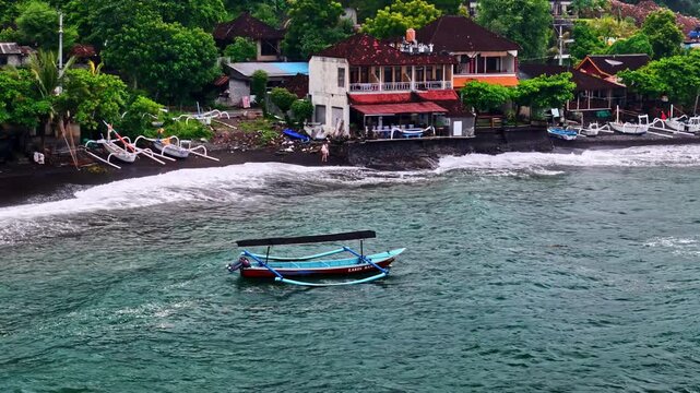 Aerial view of Amed Beach with traditional outrigger boats on black sand, coastal buildings, and turquoise ocean waves in Bali, Indonesia.