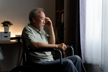 Fototapeta premium Stressed elderly man sitting in wheelchair at home, holding glasses and pressing his forehead with closed eyes, showing discomfort, emotional strain, exhaustion and challenges of aging alone.