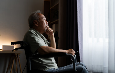Lonely older man sitting in a wheelchair beside a window, resting his chin on his hand and looking...