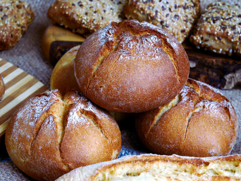 A close-up shot features several rustic loaves of bread, some dusted with flour and others topped with seeds. The bread appears golden brown and freshly baked, arranged on a textured surface.