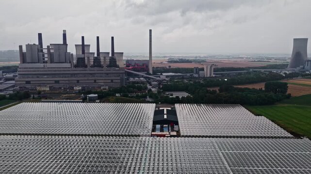 Aerial view of RWE Power AG Kraftwerk Neurath power plant with cooling towers and adjacent large greenhouse complex under a cloudy sky in Grevenbroich, Nordrhein-Westfalen, Germany.