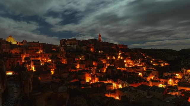 Night time lapse of moving clouds and stars over illuminated Sassi. Ancient old town of Matera in Italy.