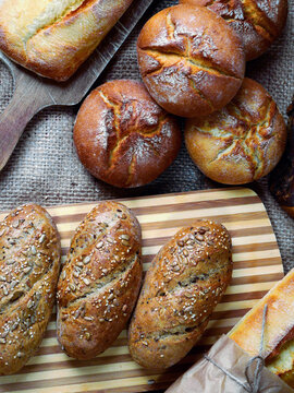 A top-down view of an assortment of freshly baked breads, including several round rolls, elongated loaves, and baguettes, are arranged on a rustic surface with burlap and wooden boards.