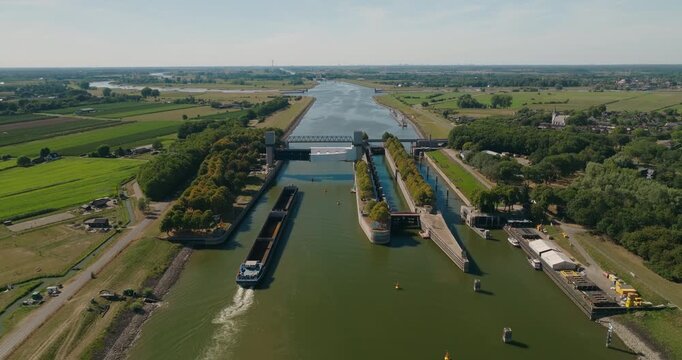 Wide aerial view of a Dutch river sluice complex with a barge passing beneath an open upward gate, surrounded by vast green polder and agricultural landscape under blue summer skies.