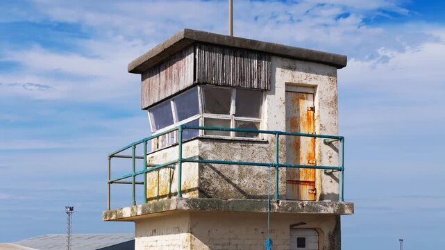 Old disused watchtower at Ayr Harbour, Scotland, showing weathered concrete, faded timber cladding and metal railings against a blue sky, cinematic aerial orbital view