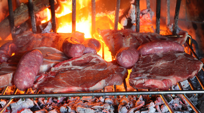 Raw meat assortment with sausages and pork steaks cooking on grill over fireplace wood embers