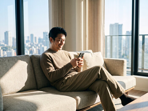 Smiling middle‑aged Japanese man sits comfortably on living‑room sofa, happily browsing content on his smartphone by large balcony windows.