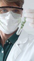 Adult male scientist in mask and gloves holding test tube with green plant sample, conducting...