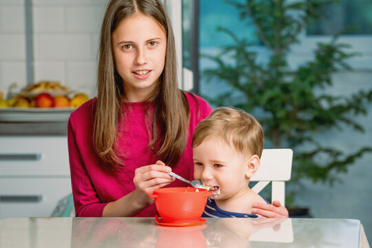 Older sister feeding her little brother with a spoon at home.