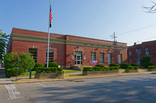 Small-town United States post office exterior with American flag on a flagpole, red brick facade, manicured shrubs and a sunny blue sky, Editorial Use only Plymouth, IN USA Aug 21, 2026. Conveying civ