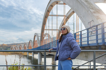 Adult female traveler poses confidently beside the bridge over Jezioro Mucharskie, wearing a blue puffer jacket, matching joggers, knitted beanie and sunglasses. Strong architectural perspective and a © Volha
