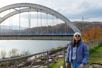 Adult woman wearing sunglasses, knitted beanie and blue puffer jacket stands beside Jezioro Mucharskie with the distinctive concrete arched bridge behind her. Autumn forest and mountain scenery create © Volha