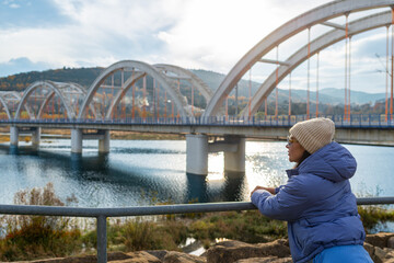 Female traveler in warm blue clothing leans on a railing and looks toward the repeating concrete arches crossing Jezioro Mucharskie. Autumn mountains, reflective water and soft sunlight create a conte © Volha