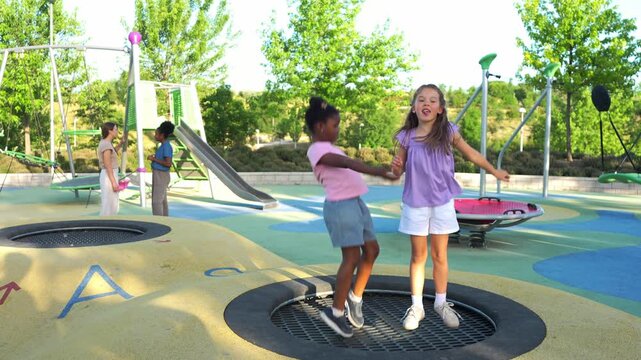 Two cheerful multiethnic girls, an african american and a caucasian, happily jumping on an in ground trampoline at a sunny park playground