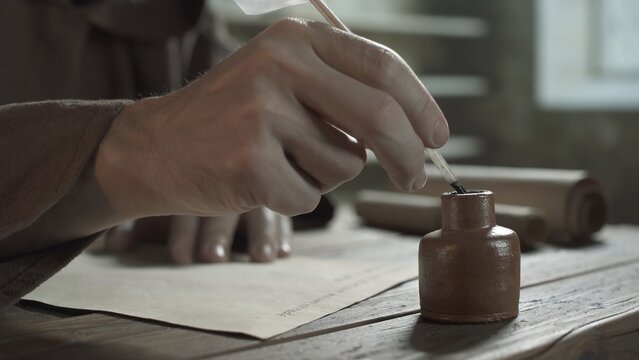 Hand of a monk dipping a quill pen into a ceramic inkwell on a rustic wooden table inside a medieval monastery scriptorium