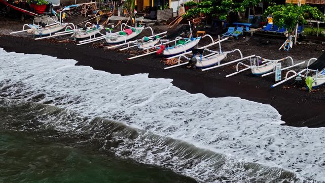 Aerial view of traditional outrigger boats resting on the black sand shore of Amed Beach with crashing waves in Bali, Indonesia.