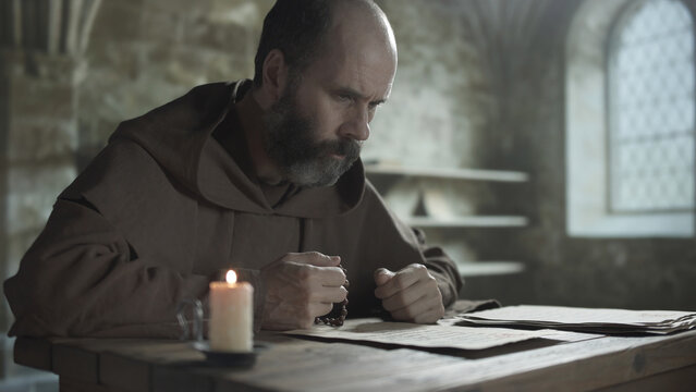 Monk holding wooden rosary beads and praying over a manuscript sheet at a wooden table near a burning candle inside a medieval scriptorium