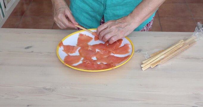 Wrapping Smoked Salmon Around Breadsticks to Prepare Savory Finger Food. Female hands wrapping slices of smoked salmon around breadsticks and arranging them on a plate to prepare a savory appetizer.