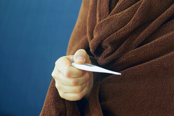 Close-up of patient sitting wrapped in blanket, holding digital thermometer in hand. Reflects home...