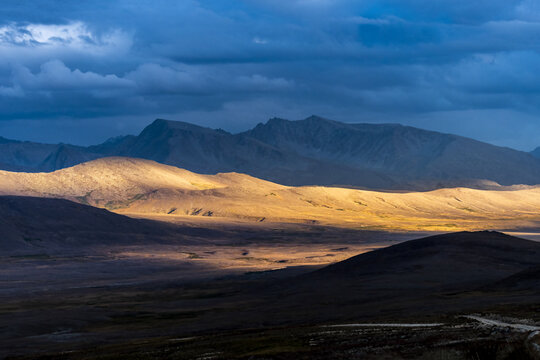 View of the vast, rolling plains of Deosai National Park illuminated by golden hour light against a backdrop of dramatic, dark blue storm clouds over the mountains in Skardu, Pakistan.