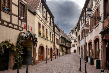 Fototapeta premium Thick garland of greenery and hanging ornaments arches over a doorway on a Kaysersberg street where sandstone arcades and shuttered facades run away under a heavy grey sky.