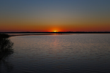 Obraz premium Beautiful sun setting behind forest horizon close up. Crimson orange sky twilight reflection on calm river water surface. Horizontal outdoor nature camping and fishing concept with copy space